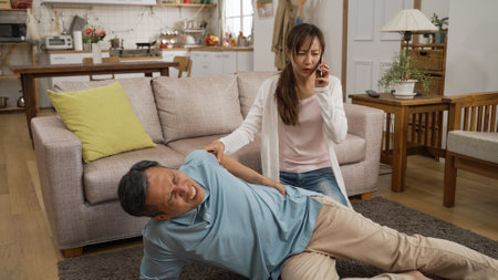 Asian woman granddaughter rushing into living room to assist fallen grandfather at home. She takes out phone and calls 911 immediately as he is letting out painful cryの写真素材