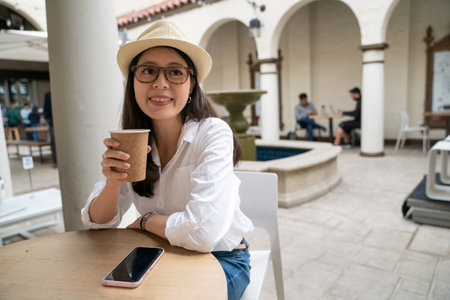 cheerful asian chinese girl visitor wearing hat having hot drink and enjoying her stay at a local coffee shop while visiting palo alto city California usaの写真素材