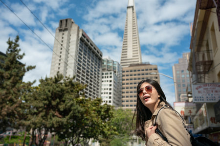 amazed asian Taiwanese woman tourist admiring streetscape with Transamerica Pyramid looming up among tall buildings against blue sky at background in san Francisco's Chinatownの写真素材