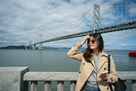 smiling asian Taiwanese female traveler enjoying beautiful view of skyline and Oakland bay bridge at san Francisco bay while relaxing by outdoor railing at ferry buildingの写真素材