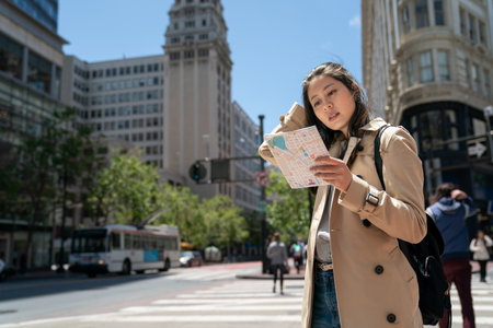 confused asian woman traveler pushing hair back and studying route map with concentration on the street while visiting downtown san Francisco in California usa on breezy dayの写真素材
