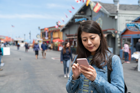 smiling asian Japanese woman traveler looking for local seafood restaurant to try online with phone during her visit at Old Fisherman's Wharf in California usaの写真素材