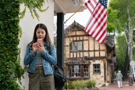 asian woman tourist searching for online guide on smartphone by a building with American flag at background during her spring trip in Carmel by the sea the coastal townの写真素材