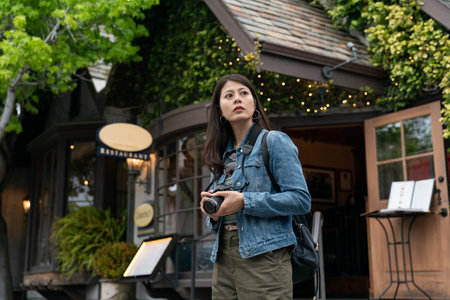 asian chinese woman holding camera and looking into space with curiosity while exploring Carmel by the sea near a wooden cottage style restaurant in usaの写真素材