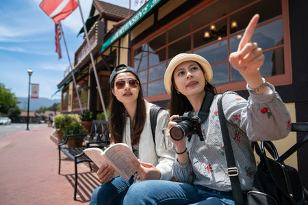 Asians taiwanese female photographer pointing finger at popular attraction in distance as her friend is consulting travel book on outdoor bench at sidewalk in downtown solvang californiaの写真素材