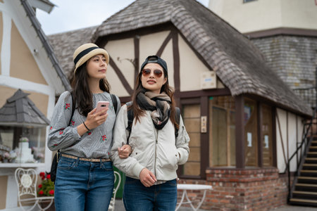 two happy asian Korean girls looking at charming Danish rural house buildings on the street while using phone for travel info during their stay in solvang California usaの写真素材