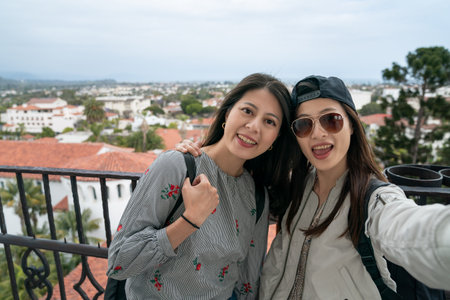 two smiling asian chinese female friends looking at camera together while taking selfie picture with beautiful clock tower view backdrop on balcony of santa Barbara county courthouse in Californiaの写真素材