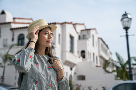 smiling young asian chinese woman visitor holding hat and looking at streetscape in downtown santa...