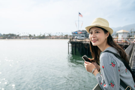 solo travel lifestyle and people on vacation in usa. smiling asian korean young female visitor admiring pretty coastal scenery at waterfront railing at santa Barbara harbor in californiaの写真素材