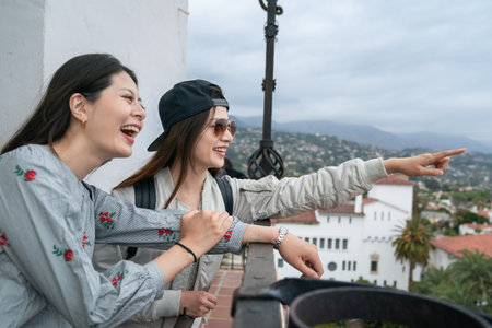 asian Japanese girl wearing cap pointing finger at popular attraction in distance to her smiling friends as they are looking at city view from balcony at santa Barbara courthouseの写真素材