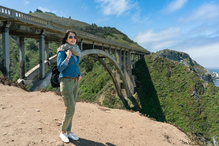 full length of fashionable asian Korean girl admiring natural beauty with backdrop of big sur bridge in California usaの写真素材