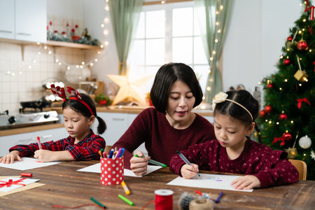 asian schoolgirls writing letters to Santa Claus with the help of their parent in a beautiful decorated room for Christmas and new year on backgroundの写真素材