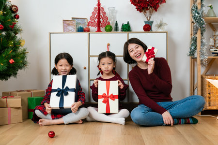 happy asian mother and her young daughters showing Christmas gifts while smiling at the camera in a cozy living room with Christmas decorationsの写真素材