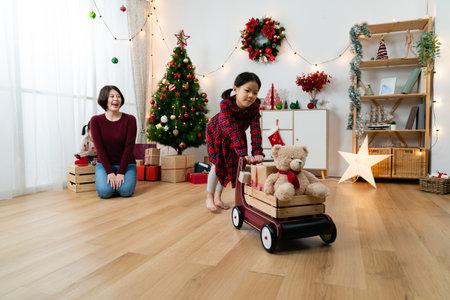 happy Korean girl is having fun pushing a toy wagon in a modern living room with Christmas decoration while her mother is smiling at her behindの写真素材