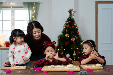 asian schoolgirl carving her dough with a mold while her mother and sisters are all looking at her at table. family baking fun for Christmas holiday at homeの写真素材