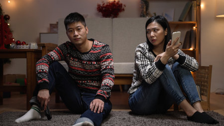 unhappy asian married couple pushing each other while they are sitting on the ground watching tv and using phone after a fight in the living room at homeの写真素材