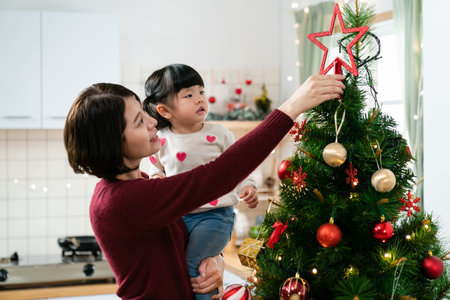 happy asian mother holding her cute baby daughter and fixing the star topper on the Christmas tree while preparing for xmas winter holiday at homeの写真素材
