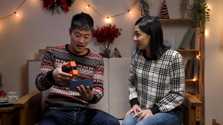 woman gesturing at empty area and surprising her husband with present. smiling asian man showing surprise expression while opening gift in a christmas living roomの写真素材