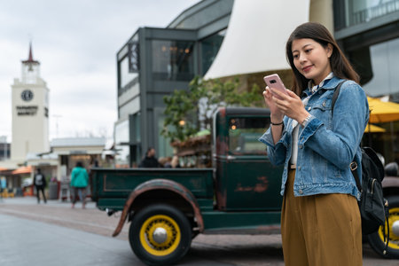 Cheerful young Asian woman wearing coat walking outdoors using mobile phone in front of farmers market in Los Angeles, USA.の写真素材