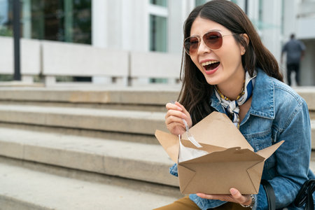 charming Asian girl sits leisurely on the steps and laughs while eating lunch in Los Angeles, USAの写真素材
