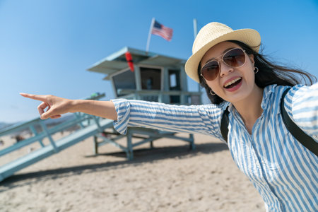 excited asian chinese woman traveler looking at camera and showing something fun in distance with pointing gesture near Lifeguard Tower on santa monica beachの写真素材