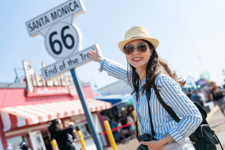 dutch angle of happy stylish asian Japanese woman visitor smiling at camera and showing route 66 end of the trail sign at background with hand gestureの写真素材