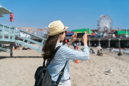 rear view of asian Japanese girl backpacker taking photos with smartphone on beach of santa monica pier near Lifeguard Towerの写真素材