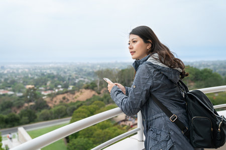 smiling asian Chinese girl backpacker leaning on the balcony railing of a building to enjoy cityscape of los angeles while traveling in united statesの写真素材