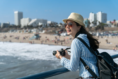happy asian taiwanese girl backpacker holding camera and leaning on waterfront railing while looking at ocean scenery at beach of santa monica pierの写真素材