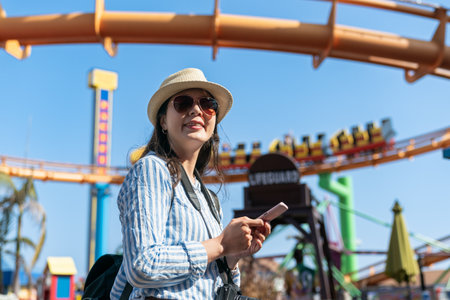smiling asian Chinese woman visitor holding phone standing under exciting roller coaster ride while having fun visiting santa monica amusement park in California usaの写真素材