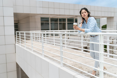 full length of asian Japanese business lady drinking coffee and leaning against bridge railing to relax with urban background in the city of los angeles usaの写真素材