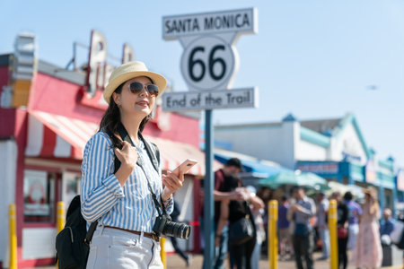 fashionable asian Chinese female photographer with phone enjoying sunny weather and townscape near famous landmark the end of route 66 sign at santa monica pierの写真素材
