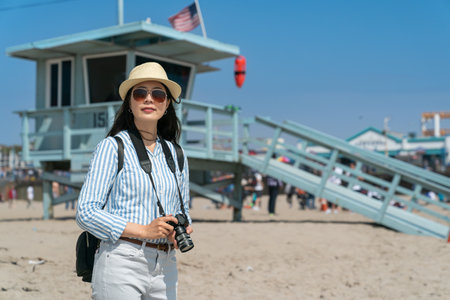 fashionable asian Korean girl photographer on vacation looking at beautiful scenery at santa monica beach with lifeguard tower at backgroundの写真素材