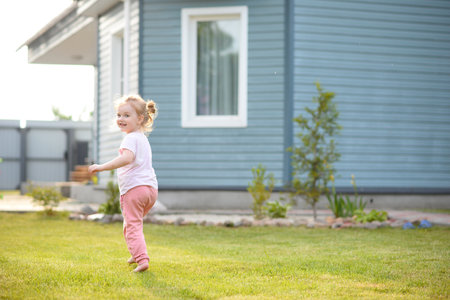 A small child runs on a green lawn. A beautiful and cheerful girl plays on the lawn near the houseの写真素材