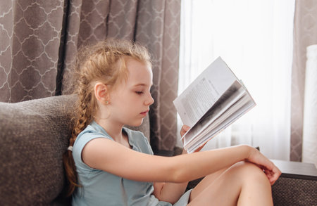 Girl reading a book for school program sitting on the couch. The theme of books and reading.