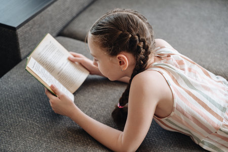 A girl comfortably lying on a sofa, engrossed in reading a book, depicting relaxationの写真素材