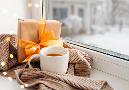 Cozy winter still life with mug, gift box, and scarf on windowsill, snowy courtyard outsideの写真素材