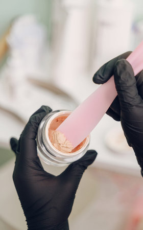 Hands in black gloves mixing pink cosmetic cream in jar, skincare laboratory process.の写真素材