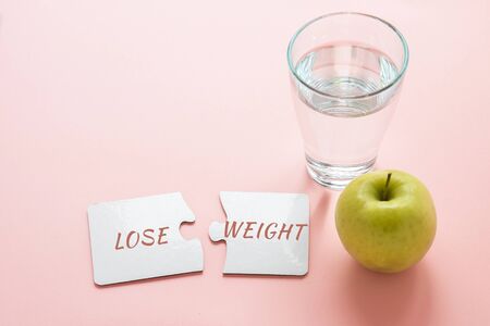 two white disconnected puzzles with the inscription lose weight, a glass of water and a green apple on a pink background, diet conceptの写真素材