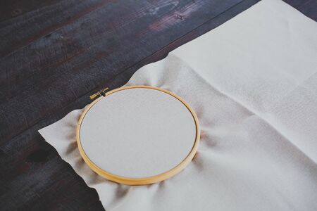 Canvas for embroidery with a cross embroidered in a round wooden hoop lies on a dark table
