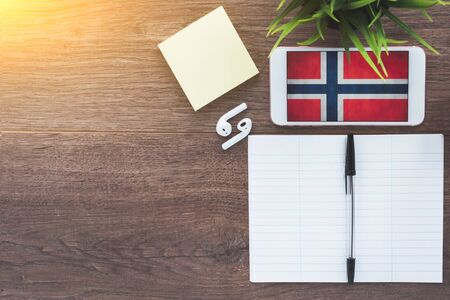 norwegian flag smartphone, wireless headphones, notepad, green plant on brown desktop, place for textの写真素材
