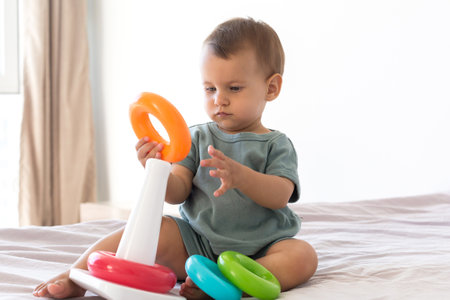 A small infant sits on a bed playing with a multi-colored plastic stacking toy