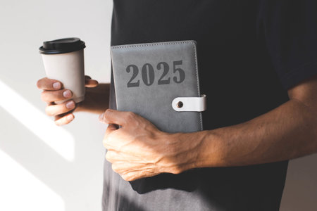 male hand holding a diary with the text 2025 and paper cup with hot drink on the background of a black T-shirt, concept of planning goals and plans for the new year.の写真素材
