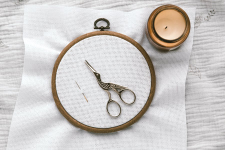 Embroidery hoop with white canvas, vintage stork-shaped scissors and needle on textured fabric, next to a lit candle in warm cozy atmosphere, representing creativity and handmade crafts, cross stitchの写真素材