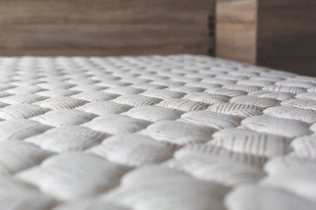 Close-up of white textured mattress surface with soft quilted pattern, in cozy bedroom interior with wooden furniture, concept of comfort, sleep and relaxationの写真素材