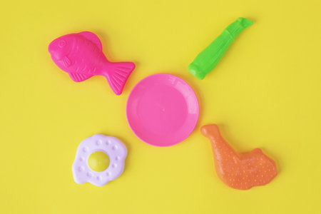 Colorful plastic toy food items, including a fish, chicken leg, egg, plate, and vegetable, arranged on a bright yellow background, symbolizing children's play and imaginationの写真素材