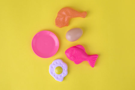 Colorful plastic toy food items, including a fish, chicken leg, egg, plate, and donut, arranged on a bright yellow background, representing children's play and imaginationの写真素材