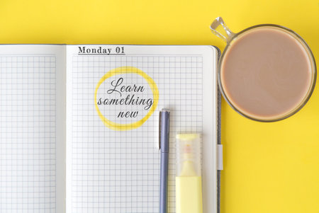 Open notebook with grid pages, handwritten quote text learn something new highlighted in yellow, pen, marker and cup of coffee on bright yellow background, concept of education and motivationの写真素材