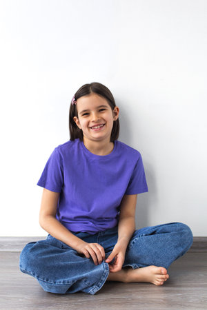 Smiling white brunette girl aged 7-8 in purple t-shirt and jeans sitting barefoot on wooden floor against white wall.の写真素材