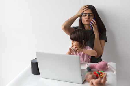 Stressed mother working remotely on a laptop while talking on the phone and caring for her child playing with toys at the same tableの写真素材